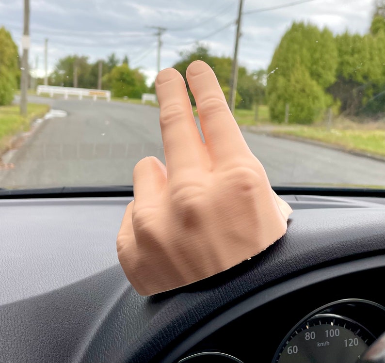 May include: A 3D printed hand model with two fingers extended in a peace sign gesture. The hand is resting on the dashboard of a car.