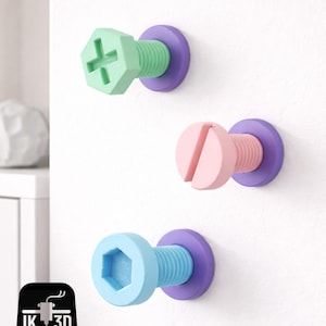 Puede incluir: Tres ganchos de pared con forma de tornillo de colores. Los ganchos tienen una cabeza verde, rosa y azul con una base morada. Las roscas de los tornillos son del mismo color que la cabeza. Los ganchos están montados en una pared blanca.