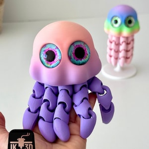 May include: A 3D-printed, articulated jellyfish toy in shades of pink, purple, and blue. The toy has large, detailed eyes and jointed tentacles. A smaller, rainbow-colored jellyfish is in the background. The logo "IK 3D" is in the lower left corner.