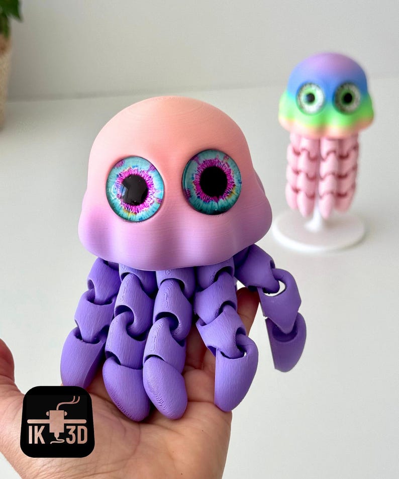 May include: A 3D-printed, articulated jellyfish toy in shades of pink, purple, and blue. The toy has large, detailed eyes and jointed tentacles. A smaller, rainbow-colored jellyfish is in the background. The logo "IK 3D" is in the lower left corner.