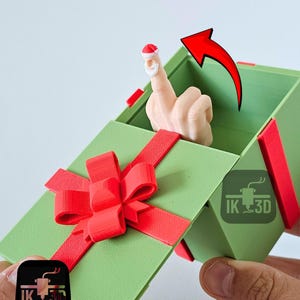Pode incluir: Uma caixa de presente verde com laço e fita vermelhos, revelando uma mão com um dedo usando um gorro de Papai Noel. A imagem inclui uma seta vermelha e o logotipo "IK 3D".