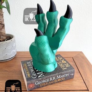 May include: A green dragon claw bookend with black claws. The bookend is holding up a copy of "Fire and Blood" by George R.R. Martin.