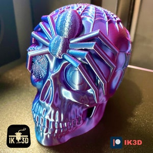 May include: A 3D printed skull with a spider on top. The skull is a deep purple and blue color with a metallic sheen. The spider is a light blue color with a metallic sheen. The spider has a web design on its back.