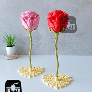 Puede incluir: Dos esculturas decorativas de rosas, una rosa y otra roja, cada una con un dise&ntilde;o de calavera en el centro de la flor. Los tallos son verdes y espinosos, y las bases son formas de telara&ntilde;a amarillas. Una peque&ntilde;a planta en maceta est&aacute; en el fondo.