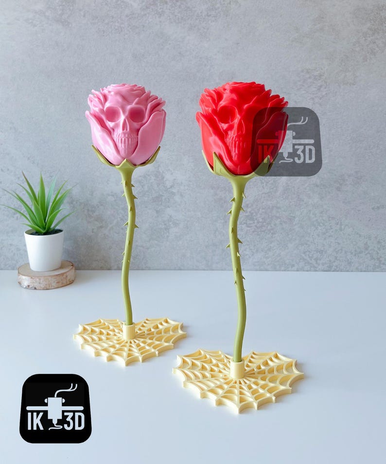 Puede incluir: Dos esculturas decorativas de rosas, una rosa y otra roja, cada una con un dise&ntilde;o de calavera en el centro de la flor. Los tallos son verdes y espinosos, y las bases son formas de telara&ntilde;a amarillas. Una peque&ntilde;a planta en maceta est&aacute; en el fondo.
