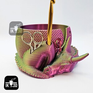 May include: A 3D printed yarn bowl in the shape of a sleeping dragon with a pink and green iridescent finish. The bowl has a hole for a crochet hook and a slot for scissors.