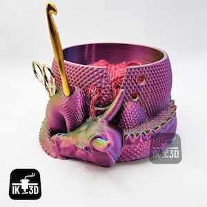 May include: A purple and green iridescent dragon yarn bowl with a gold crochet hook and scissors. The bowl has a textured surface and is designed to hold yarn and other knitting supplies. The dragon is curled up with its head resting on the bowl. The bowl is labeled "IK 3D".