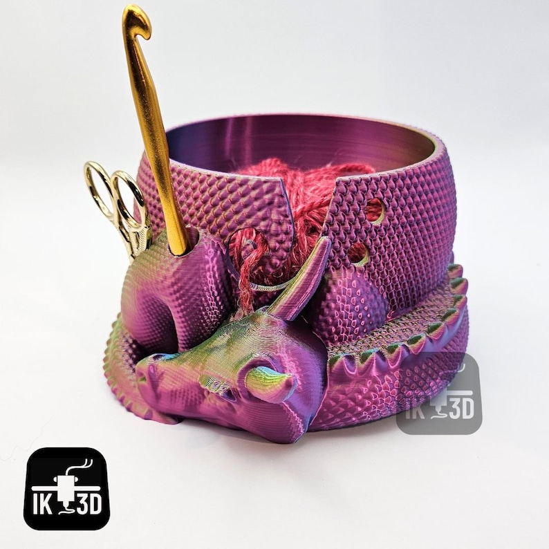 May include: A purple and green iridescent dragon yarn bowl with a gold crochet hook and scissors. The bowl has a textured surface and is designed to hold yarn and other knitting supplies. The dragon is curled up with its head resting on the bowl. The bowl is labeled "IK 3D".