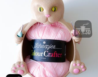 May include: A beige cat-shaped yarn holder with a pink nose and yellow eyes, holding a ball of pink yarn labeled "Scheepjes Colour Crafter." The holder has paw-shaped feet and holds two crochet hooks.