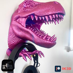 May include: A pink and purple dinosaur head wall mount with a black headphone holder. The dinosaur's mouth is open and its teeth are visible. The holder is attached to the dinosaur's jaw. The text "IK3D" is visible on the holder.