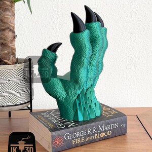May include: A green dragon claw bookend with black claws, resting on a book titled "Fire and Blood" by George R.R. Martin.