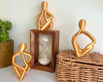 May include: Three gold-colored abstract sculptures of people sitting in a contemplative pose. The sculptures are arranged on a wooden shelf with an hourglass in the middle. A wicker basket is on the right side of the shelf.