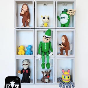 May include: A display of nine figurines in individual white cubbies. The figurines include a brown Bigfoot giving a thumbs up, a green leprechaun holding a sign that says "Kiss me I'm Irish", and a green elf. Other figurines include a yellow dinosaur and a brown reindeer.