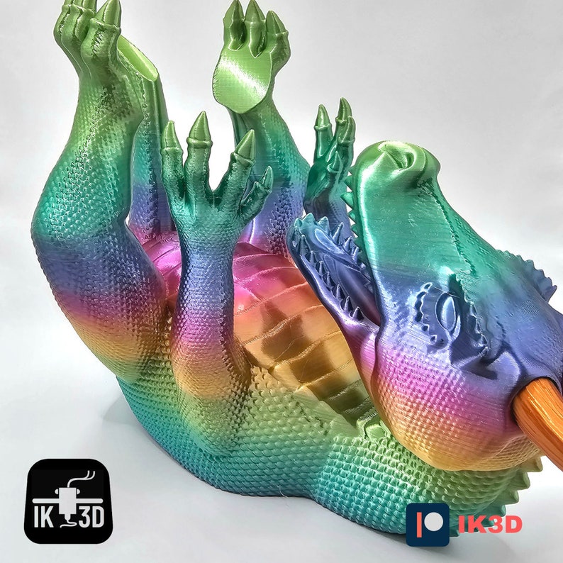 May include: A rainbow iridescent 3D printed dragon figurine with an open mouth, holding a wooden dowel in its mouth. The dragon is lying on its back with its legs in the air. The figurine is made by IK3D.