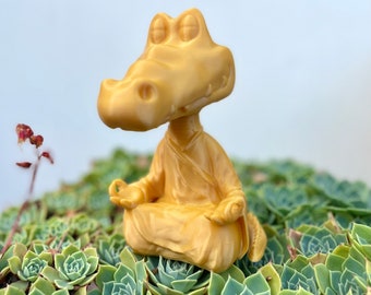 May include: A golden figurine of a crocodile meditating in a lotus position. The crocodile is wearing a robe and has its eyes closed. The figurine is sitting on a bed of green succulents.