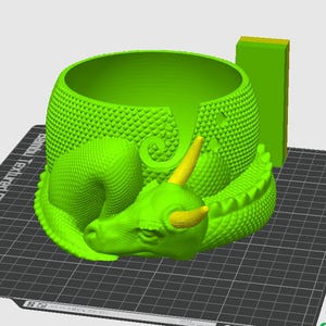 May include: A green 3D printed dragon yarn bowl with a textured surface. The bowl is shaped like a sleeping dragon with a curled tail and horns. The bowl is sitting on a black grid surface.