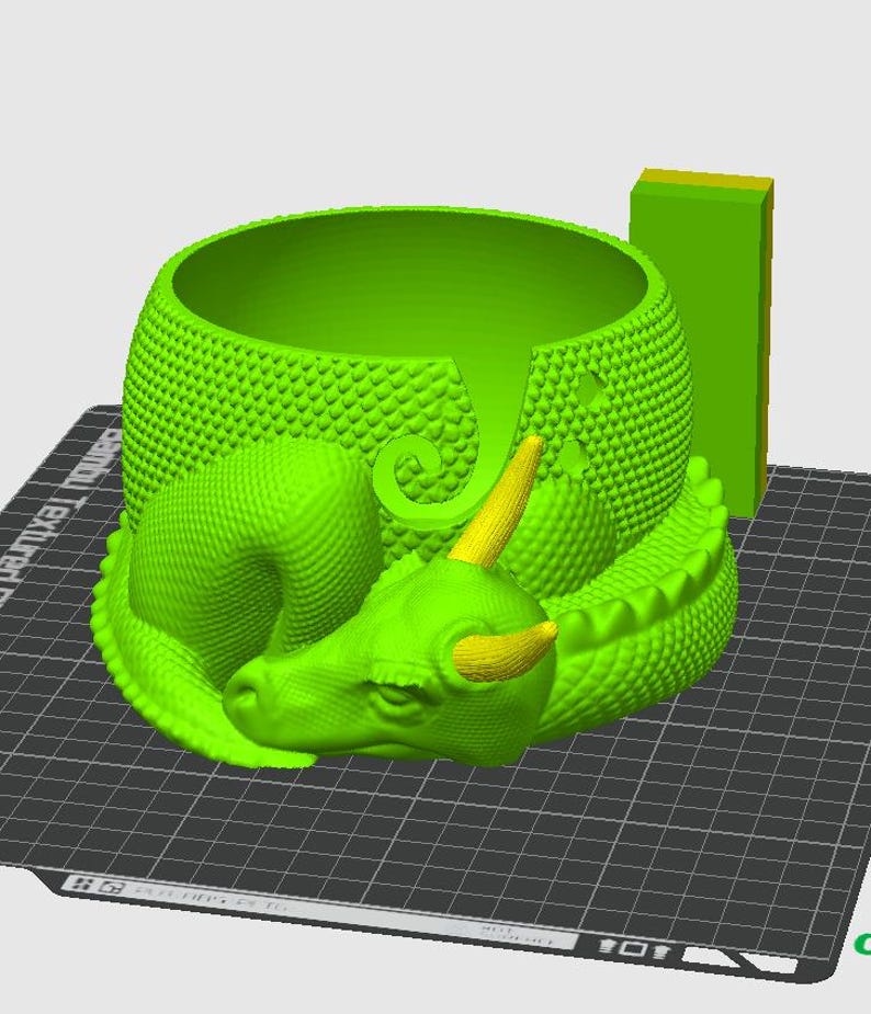 May include: A green 3D printed dragon yarn bowl with a textured surface. The bowl is shaped like a sleeping dragon with a curled tail and horns. The bowl is sitting on a black grid surface.