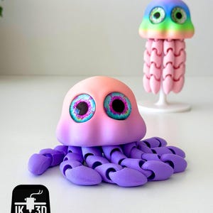 May include: Two 3D-printed jellyfish toys. The larger one is pink and purple with large, detailed eyes. The smaller one is rainbow-colored with pink tentacles. The toys are on a white surface. The logo "IK 3D" is in the bottom left corner.