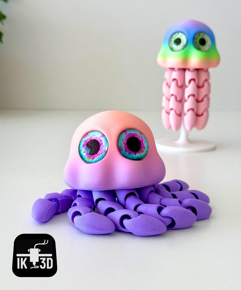 May include: Two 3D-printed jellyfish toys. The larger one is pink and purple with large, detailed eyes. The smaller one is rainbow-colored with pink tentacles. The toys are on a white surface. The logo "IK 3D" is in the bottom left corner.