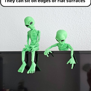 Puede incluir: Dos figuritas de extraterrestres verdes sentadas en una superficie negra. El extraterrestre de la izquierda est&aacute; sentado con las piernas cruzadas. El extraterrestre de la derecha est&aacute; inclinado hacia adelante con los brazos extendidos. El texto "They can sit on edges or flat surfaces" se muestra sobre los extraterrestres.