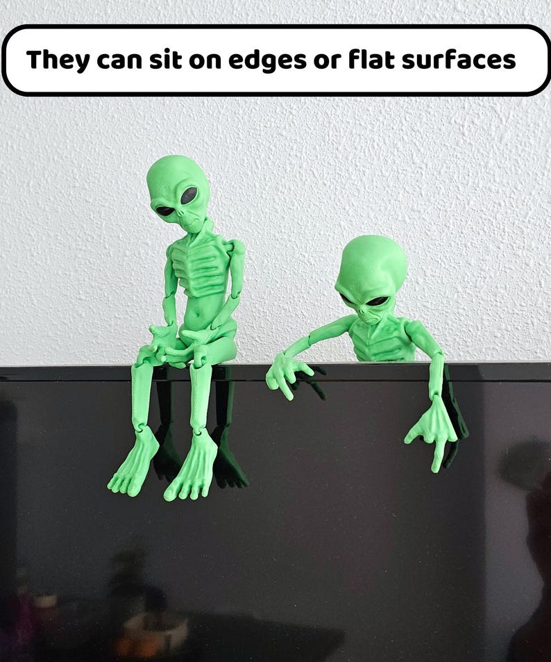 Puede incluir: Dos figuritas de extraterrestres verdes sentadas en una superficie negra. El extraterrestre de la izquierda est&aacute; sentado con las piernas cruzadas. El extraterrestre de la derecha est&aacute; inclinado hacia adelante con los brazos extendidos. El texto "They can sit on edges or flat surfaces" se muestra sobre los extraterrestres.