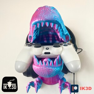May include: A pink and blue 3D printed dinosaur head with an open mouth, holding a white video game controller and black headphones. The dinosaur is a wall mount for headphones and controllers. The text "IK3D" is on the bottom left and right of the image.