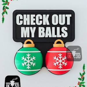 Pode incluir: Uma placa preta com o texto "CHECK OUT MY BALLS" acima de duas decorações de enfeites de Natal. Um enfeite é verde, o outro vermelho, ambos com desenhos de flocos de neve brancos. Os enfeites têm tampas douradas.