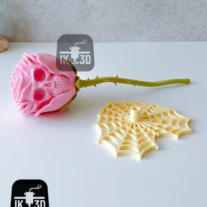 Puede incluir: Una rosa rosa con una flor en forma de calavera y un tallo verde con espinas. Junto a ella, un soporte amarillo con forma de telara&ntilde;a. La rosa es un art&iacute;culo decorativo &uacute;nico.