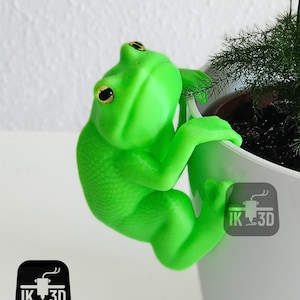 May include: A bright green frog figurine climbing on a white plant pot. The frog has large, golden eyes and is textured to resemble skin. The pot contains a small green plant. The logo "IK 3D" is visible.