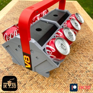 May include: A gray and red 3D printed can holder with the text "V8" printed on the side. The holder is designed to hold six cans and has a handle on top. The holder is sitting on a woven surface.