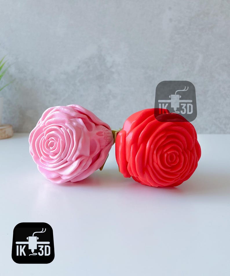 Puede incluir: Dos esculturas de rosas impresas en 3D, una rosa y otra roja, se exhiben sobre una superficie blanca. Las rosas tienen dise&ntilde;os detallados de p&eacute;talos y est&aacute;n conectadas por un tallo verde. El logotipo "IK 3D" es visible.