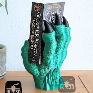 Op de afbeelding: Een groene drakenklauw boekenlegger met zwarte klauwen die een boek vasthouden met de titel "Fire and Blood" van George R.R. Martin. De boekenlegger is gemaakt van 3D-geprint plastic.