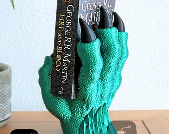May include: A green dragon claw bookend with black claws holding a book titled "Fire and Blood" by George R.R. Martin. The bookend is made of 3D printed plastic.