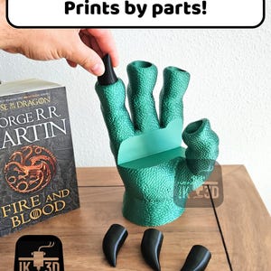 May include: A green 3D printed hand with four fingers and a thumb, with the text "Prints by parts!" above it. The hand is holding four black 3D printed claws. The hand is sitting on a wooden surface.