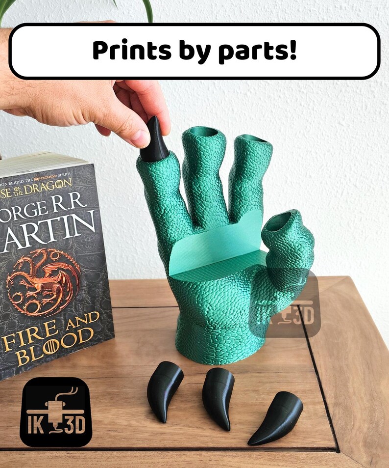 May include: A green 3D printed hand with four fingers and a thumb, with the text "Prints by parts!" above it. The hand is holding four black 3D printed claws. The hand is sitting on a wooden surface.