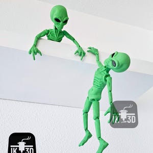 Puede incluir: Dos figuritas de extraterrestres verdes, una colgando boca abajo de una estanter&iacute;a y la otra mirando por encima del borde de la estanter&iacute;a. Ambas figuritas tienen extremidades articuladas y est&aacute;n hechas de pl&aacute;stico impreso en 3D.