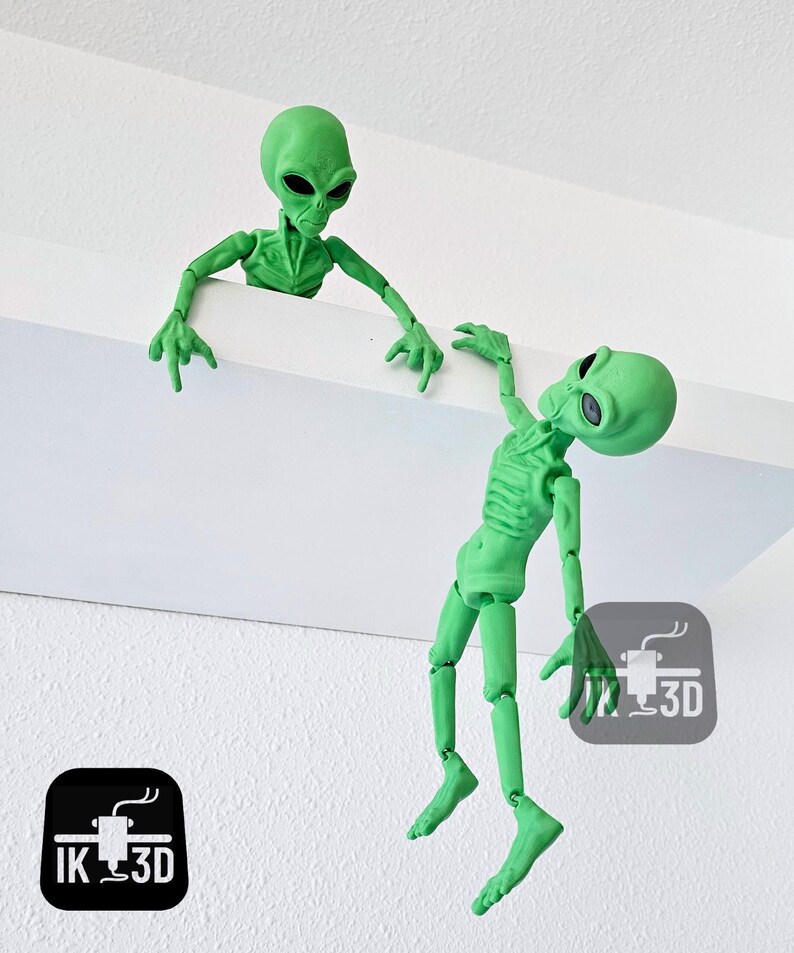 Puede incluir: Dos figuritas de extraterrestres verdes, una colgando boca abajo de una estanter&iacute;a y la otra mirando por encima del borde de la estanter&iacute;a. Ambas figuritas tienen extremidades articuladas y est&aacute;n hechas de pl&aacute;stico impreso en 3D.