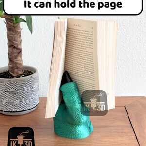May include: A green dinosaur-shaped bookend holding a book open. The bookend is made of a textured material and has a black claw. The bookend is sitting on a wooden surface.