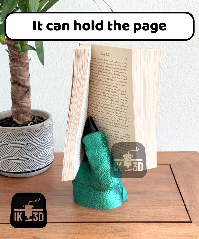 May include: A green dinosaur-shaped bookend holding a book open. The bookend is made of a textured material and has a black claw. The bookend is sitting on a wooden surface.