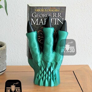 May include: A green dragon claw bookend with black claws holding a book titled "House of the Dragon" by George R.R. Martin. The bookend is on a wooden surface.