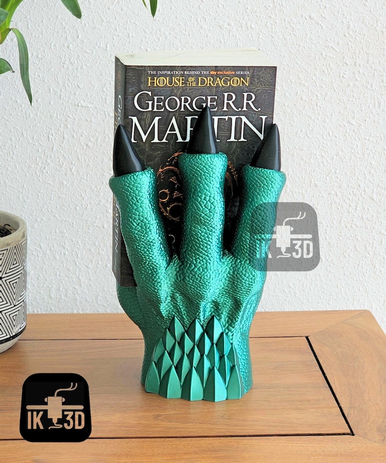 May include: A green dragon claw bookend with black claws holding a book titled "House of the Dragon" by George R.R. Martin. The bookend is on a wooden surface.
