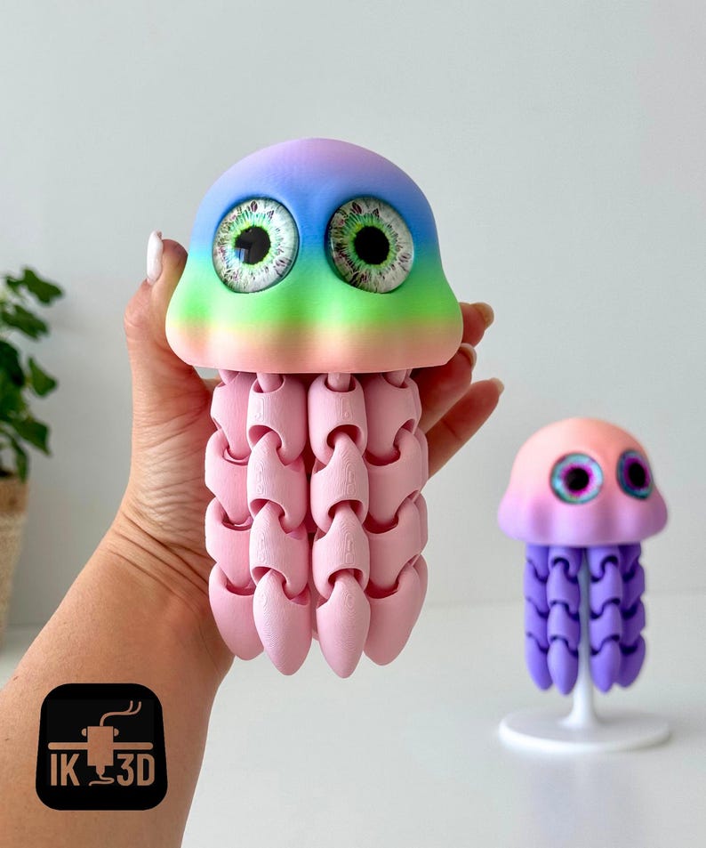 May include: A large, articulated jellyfish toy with a rainbow-colored head and pink tentacles is held in a hand. A smaller jellyfish toy with a purple body and pink head sits on a white stand. The toys have large, detailed eyes.
