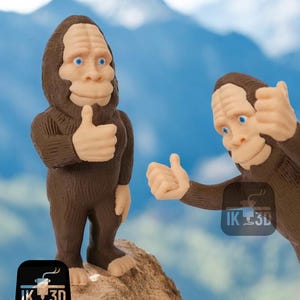 Puede incluir: Dos figuritas de Bigfoot impresas en 3D de color marrón que dan un pulgar hacia arriba. Las figuritas están de pie sobre una roca con un cielo azul y montañas en el fondo. El texto "IK 3D" es visible en la parte inferior de la imagen.