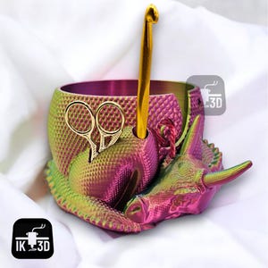 May include: A pink and green 3D printed yarn bowl shaped like a rhinoceros with a gold crochet hook and scissors. The bowl has a textured surface and a hole for the hook.