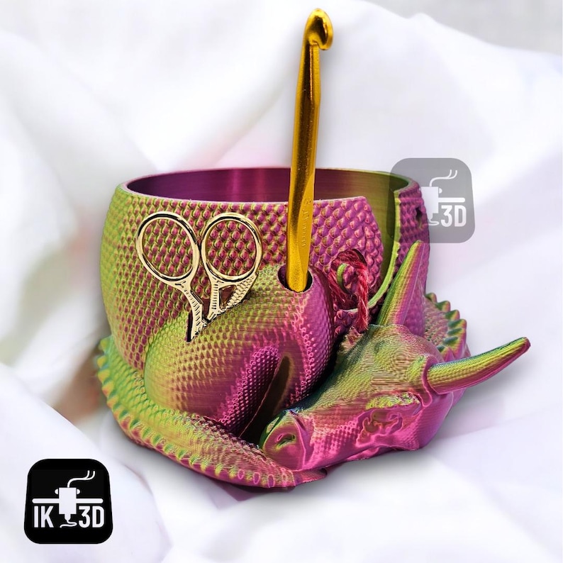 May include: A pink and green 3D printed yarn bowl shaped like a rhinoceros with a gold crochet hook and scissors. The bowl has a textured surface and a hole for the hook.
