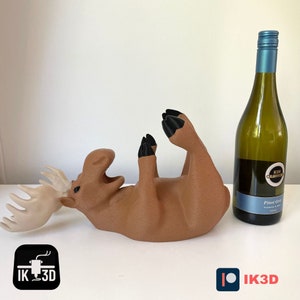 Puede incluir: Un soporte para botellas de vino en forma de alce marr&oacute;n con ojos y pezu&ntilde;as negras. El soporte est&aacute; tumbado de espaldas con las patas en el aire. Una botella de vino blanco est&aacute; al lado del soporte.