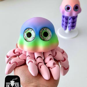 May include: A colorful, 3D-printed jellyfish toy with a rainbow-colored head and large, detailed eyes. The toy is held in a hand, with a smaller jellyfish figure in the background. The jellyfish has articulated tentacles and a pink body.
