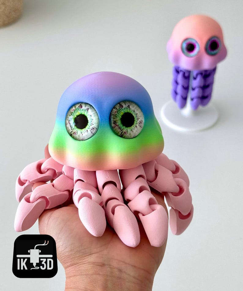 May include: A colorful, 3D-printed jellyfish toy with a rainbow-colored head and large, detailed eyes. The toy is held in a hand, with a smaller jellyfish figure in the background. The jellyfish has articulated tentacles and a pink body.