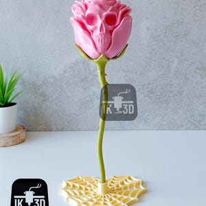 Puede incluir: Una escultura de rosa rosa con un centro en forma de calavera. El tallo es verde con espinas y la base es un dise&ntilde;o de telara&ntilde;a amarilla. La escultura tiene el texto "IK 3D" en un cuadrado negro.