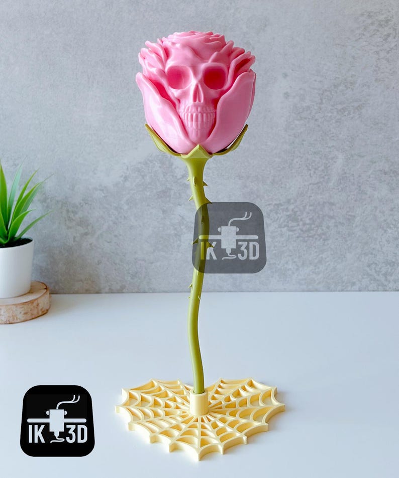 Puede incluir: Una escultura de rosa rosa con un centro en forma de calavera. El tallo es verde con espinas y la base es un dise&ntilde;o de telara&ntilde;a amarilla. La escultura tiene el texto "IK 3D" en un cuadrado negro.